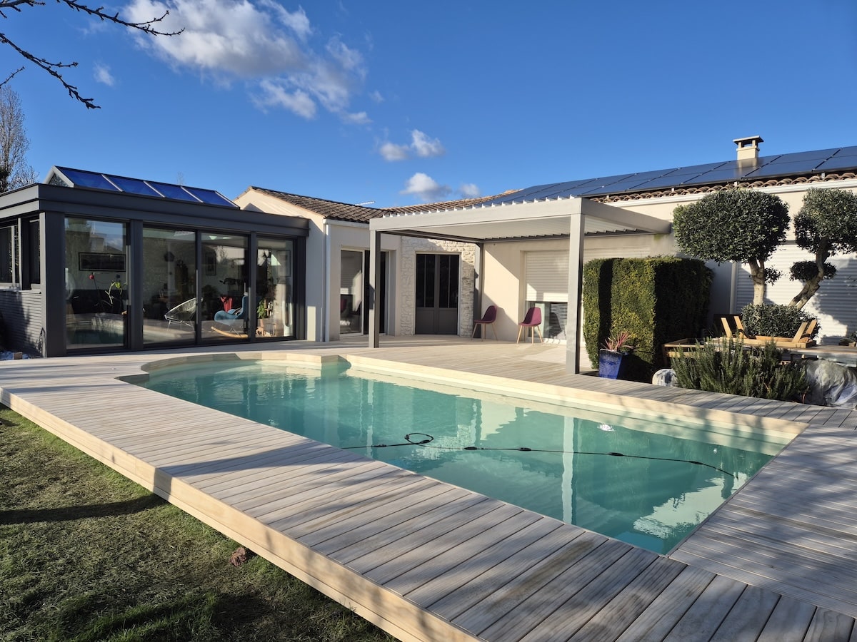Piscine rectangulaire avec terrasse en bois, véranda vitrée et pergola bioclimatique devant une maison moderne.