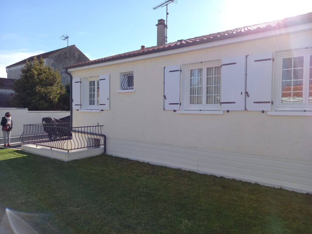 Maison blanche traditionnelle avec volets blancs, toit en tuiles et jardin ensoleillé sous un ciel bleu.